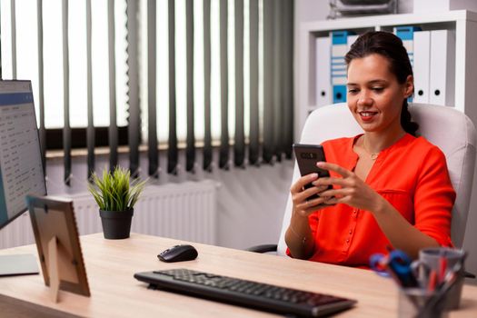 Freelance businesswoman texting corporate financial report on smartphone wearing red. Professional financial analist sitting at office desk in workplace using modern smartphone.