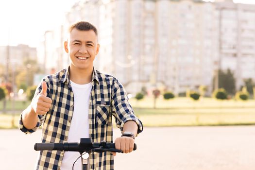 Caucasian hipster man leaning on electric scooter looking at camera cheerfully smiling and showing thumb up. Ecology and urban lifestyle. Eco-friendly modern urban transport.