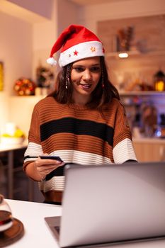 Joyful adult doing online shopping for christmas family dinner at home with seasonal decorations, lights and ornaments. Caucasian woman doing credit card payment for celebration party