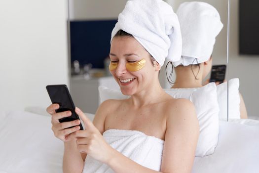 After a shower, the girl is wrapped in a towel and wears cosmetic patches for the skin under the eyes. Looks at the cell phone. Cosmetic procedures at home. Getting ready for a date.