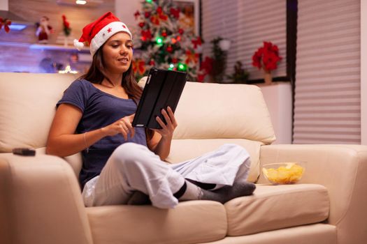 Adult sitting in lotus position on couch in xmas decorated kitchen while browsing information on internet using tablet computer. Caucasian female celebrating christmas holiday enjoying winter season