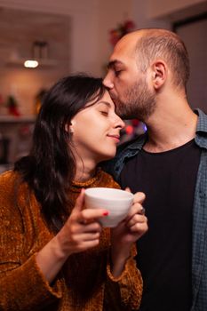 Portrait of happy couple enjoying christmas holiday spending time together in xmas decorated kitchen. Husband kissing girlfriend on forehead during winter season. New-year festive tradition