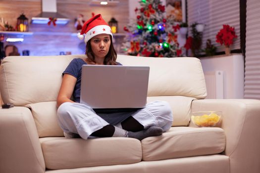 Woman writing business email on laptop sitting in lotus position on sofa during christmastime in xmas decorated kitchen. Adult with santa hat enjoying winter season celebrating christmas holiday