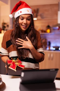 Festive young adult holding christmas gift on internet conference video call with friends and family using tablet. Caucasian cheerful woman showing present box with bow and ribbon