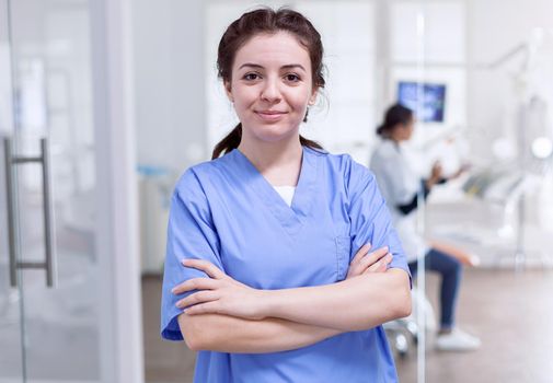 Portrait of nurse in dental reception with arms crossed looking at camera wearing uniform and dentist in the background.. Happy smiling assistant in orthodontic office.