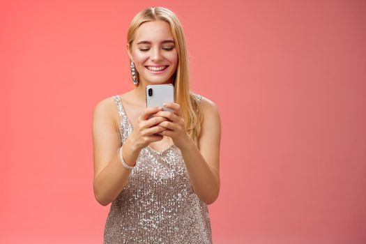 Delighted tender glamour blond woman in silver stylish glittering dress brilliand earrings holding smartphone taking photo friend capture moment celebration nightclub, red background smiling.