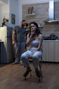 Unhappy terrified woman sitting and smoking cigarette feeling miserable after domestic violence and physical abuse from violent man. Aggressive drunk husband harassing bruised wife