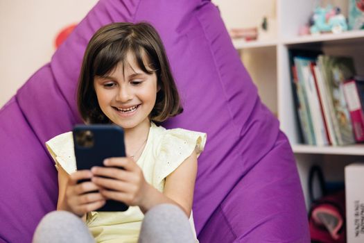 Cute caucasian kid girl holding smart phone enjoying using mobile apps, playing games at home. Small child learning in cellphone, watching video, having fun with mobile technology concept.