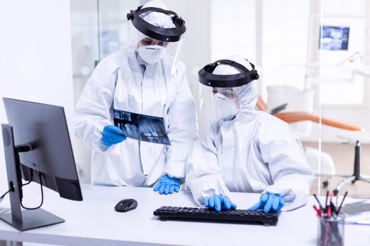 Dentist doctor and nurse in ppe suit against covid-19 and nurse holding teeth x-ray. Medical specialist wearing protective gear against coronavirus during global outbreak looking at radiography in dental office.