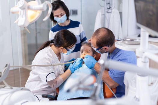 Pediatric dentist treating little girl carieswearing face mask and gloves. Mother with her kid in stomatology clinic for teeth examine using modern instruments.