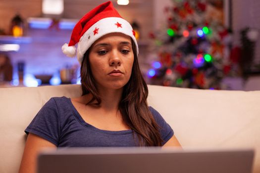 Woman looking at laptop screen reading business email during christmas holiday resting comfortable on couch in xmas decorated kitchen. Caucasian female browsing on social media. Winter season
