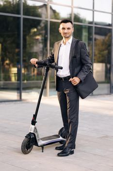 Businessman standing in front of city, holding electric scooter and looking at camera. Modern urban alternative transport. Eco-friendly transportation. Ecology and urban lifestyle.