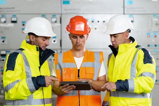 Engineers in uniform and helmets with digital tablet do pre-commissioning works inspecting electrical station in sunny premise