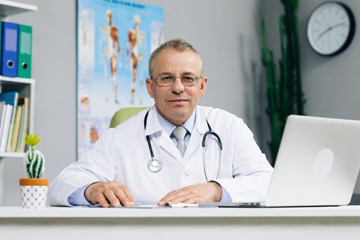 Confident old mature male head doctor physician in white medical uniform in glasses sitting at workplace. General practitioner looking at camera, posing in office.