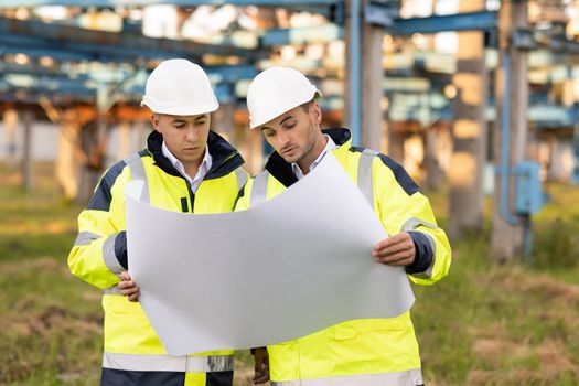 Engineers with a construction plan working near high-voltage powerline. Two engineers in special clothing discuss a drawing on paper against the background of a high-voltage power line.