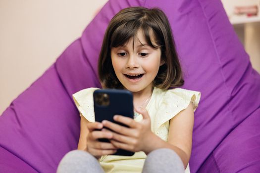 Cute child girl holds phone, kid using smartphone, child browsing the Internet, uses video communication at home on the phone.