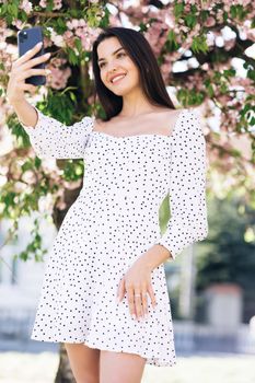 Young smiling woman in summer white dress taking selfie self portrait photos on smartphone. Model posing on park sakura trees background. Female showing positive face emotions