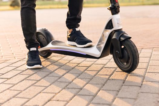 Modern Man Using Electric Scooter in Sunny Day. Adult man riding electric scooter to work. Legs man in black sneakers ride on electric mobile scooter. Eco friendly transportation.