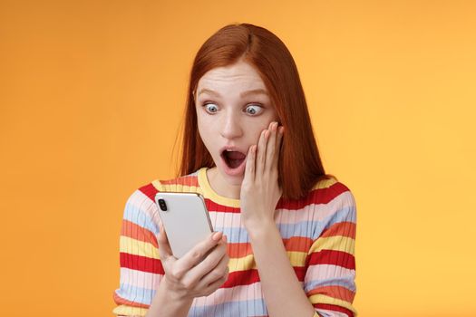 Omg what heck. Portrait shocked concerned young redhead sensitive impressed redhead woman stare smartphone display touch cheek drop jaw stunned surprised standing orange background hold phone.