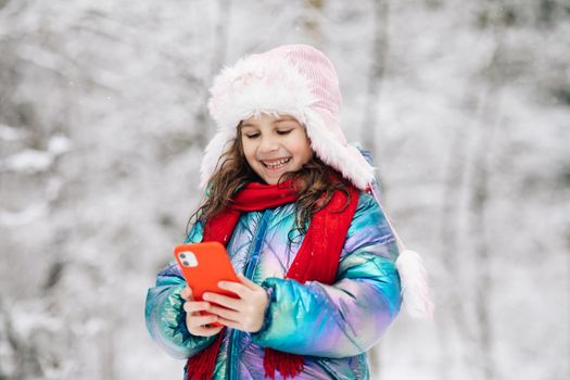 Happy little child use smartphone funny face mask app look at cell screen smiles at something funny. Holidays and celebrations concept. Outside. The dead of winter, cold weather, frost, snow