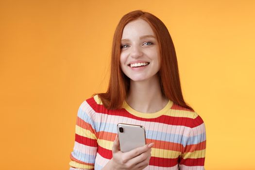 Friendly enthusiastic young redhead girl blue eyes using smartphone turn camera answer smiling broadly telling who sent message standing delighted orange background messaging, texting boyfriend.