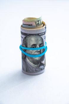 Close up photograph of a rolled up wad of cash sitting on edge with a blue rubber band wrapping across Benjamin Franklin's face on a hundred dollar bill on a white background with copy space.