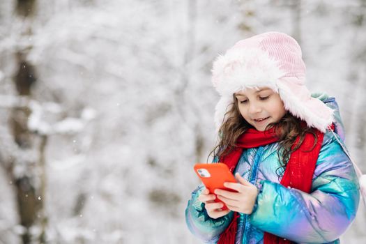 Happy little kid use smartphone funny face mask app look at cell screen smiles at something funny. Holidays and celebrations concept. Outside. The dead of winter, cold weather, frost, snow