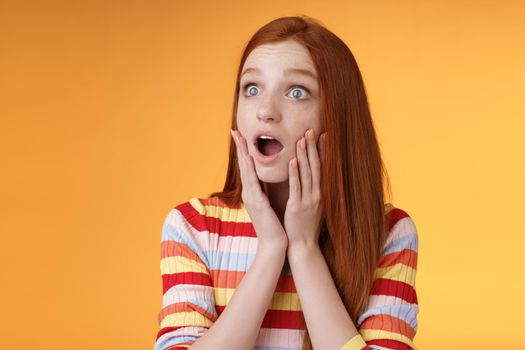 Shocked amazed young drama girl redhead blue eyes gasping empathy touch cheeks shocked drop jaw worry turn left surprised astonished standing nervously orange background. Emotions concept