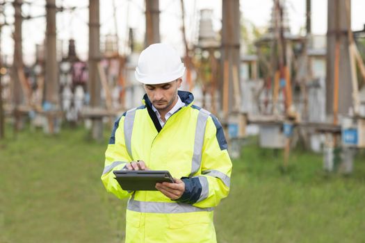 Man standing near high voltage substation and typing on device. Male engineer in uniform and helmet standing outdoor and tapping on tablet.