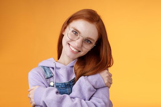 Passion, tenderness, wellbeing concept. Girl accept own self smiling charming grin tilt head hugging herself embracing body feel happiness delighted relaxing, flirty gaze camera orange background.