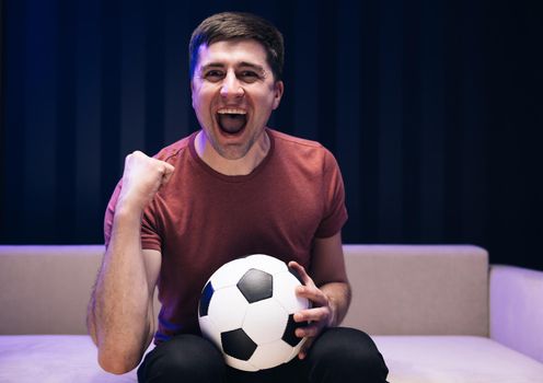 People emotions sport leisure lifestyle concept. Young fun guy 30s football fan cheer up support favorite team hold ball in red t-shirt in dark living room.