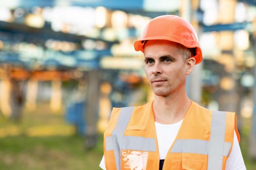 Close up portrait of male engineer in uniform and helmet standing outdoors. Man worker standing near big power electric station factory.