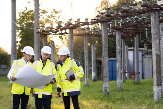 Power specialists are planning a new project outdoors. Three engineers walk near power lines in the high voltage power station. Electric industry, electrical energy production concept.