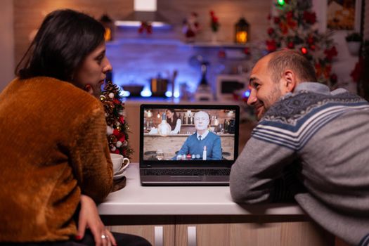 Happy family discussing with remote grandparent during online videocall conference celebrating christmas holiday together in xmas decorated kitchen. Joyful couple enjoying winter season