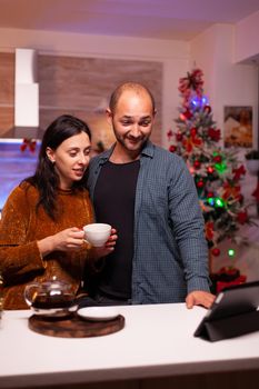 Happy couple celebrating christmas holiday with remote friends during online videocall meeting on tablet. Smiling family enjoying spending winter season time together in xmas decorated kitchen