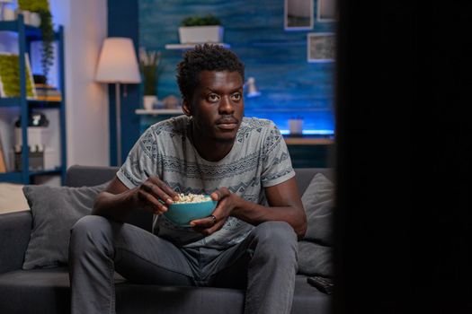 African american young man looking at football game on television resting on sofa on living room Guy watching sport channel on TV enjoying popcorn snack during free time activity at home