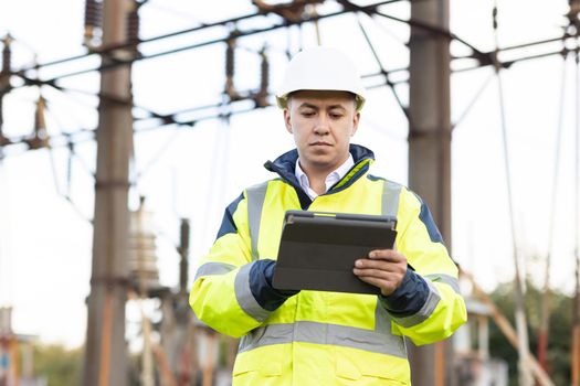 An energy engineer man in special clothes inspects a power line using data from electric sensors on a digital tablet computer. Electrical engineer with high voltage electricity pylon.
