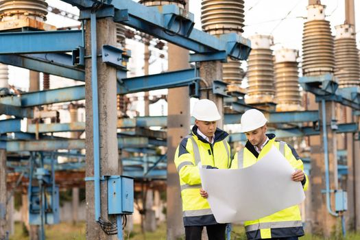 Engineers in special clothing are working on site. Two engineers discuss a drawing on paper in the open air against the background of a high-voltage power line.