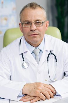 Portrait of confident old mature male head doctor physician in white medical uniform in glasses sitting at workplace. General practitioner looking at camera, posing in office.