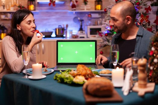 Happy family sitting at dining table in x-mas decorated kitchen enjoying winter season christmastime. Green screen mock up chroma key laptop computer with isolated display on table