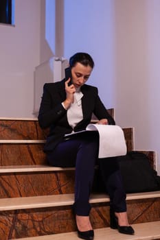 Stressed tired businesswoman readling project deadline during conversation on smartphone. Serious entrepreneur working on job project sitting on staircase of business building at night hours for job.