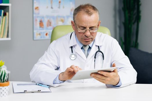 Middle aged older male doctor in white uniform holding digital computer tablet in hands, managing patients visits. Physician checking health history data in gadget, using modern app.