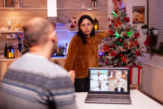 Angry nervous upset girlfriend waiting for busy boyfriend to spening time with her celebrating christmas holiday together in xmas decorated kitchen. Man having online business videocall meeting