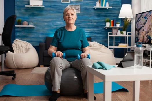 Portrait of senior woman looking into camera while sitting on swiss ball training body muscles holding fitness dumbbells during cardio gym workout. Pensioner working muscular resistance in living room