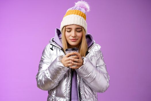 Charming tender romantic blond girl sniffing nice smell warm tasty coffee holding take-away cup warm hands close eyes look delighted wearing winter jacket hat enjoy vacation, purple background.