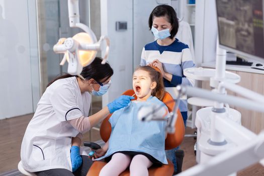 Little girl with open mouth in the course of cavity check up done by stomatologist. Dentistry specialist during child cavity consultation in stomatology office using modern technology.