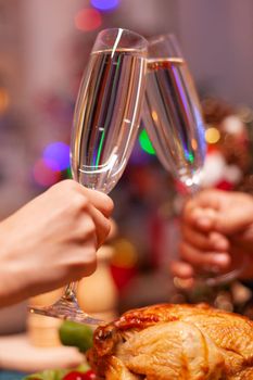 Closeup of couple hands clinking glass of wine during christmas dinner sitting at dining table in xmas decorated kitchen. Happy romantic family celebrating christmas holiday together