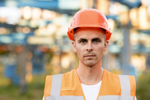 Portrait of Young Professional Heavy Industry Engineer Worker Wearing Safety Vest, Putting on Hardhat. In the Background Unfocused Large Power Station.