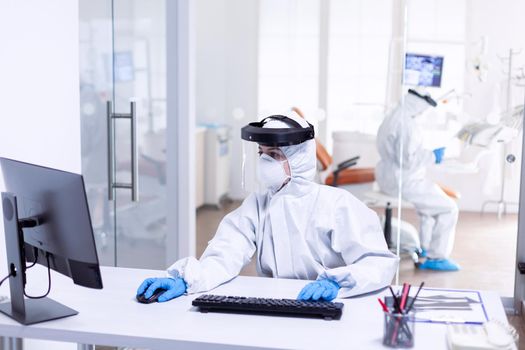 Nurse using computer during covid 19 wearing ppe suit as safety precaution. Medicine team wearing protection gear against coronavirus pandemic in dental reception as safety precaution.