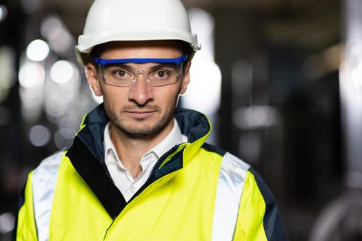 Portrait of Professional Heavy Industry Engineer or Worker Wearing Safety Uniform, Goggles and Hard Hat Looks at Camera. In the Background Unfocused Large Industrial Factory.